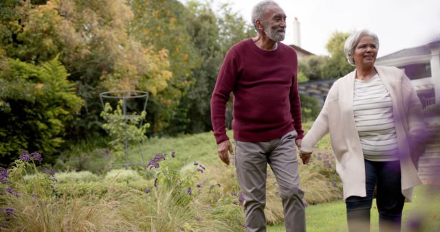 Happy diverse senior couple holding hands walking among plants in garden, copy space. Retirement, togetherness, relationship, nature and active senior lifestyle, unaltered.