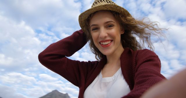 Smiling young woman with curly hair taking a selfie outdoors against a backdrop of cloudy sky. She is wearing a straw hat and a burgundy cardigan over a white top. Ideal for use in travel blogs, social media posts, tourism advertisements, or lifestyle articles showcasing joy, adventure, and natural beauty.