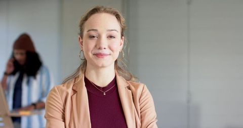 Confident young professional woman crossing arms in modern office with coworker on phone