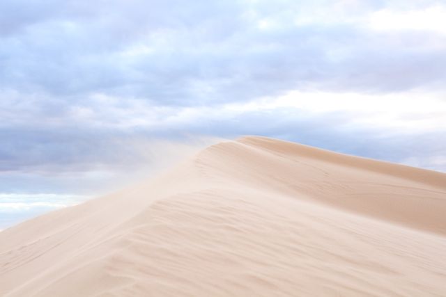 Wind blows sand from the crest of dunes under a cloudy sky, creating a serene desert landscape. Ideal for representing natural beauty, wilderness landscapes, travel destinations, and tranquility. Suitable for background images, travel brochures, nature content, and environmental websites.