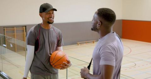 African american teammates smiling and holding basketball by indoor court glass railing