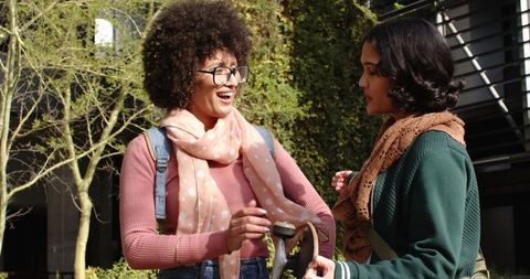 Two female students laughing and chatting while holding scooter in sunny campus courtyard