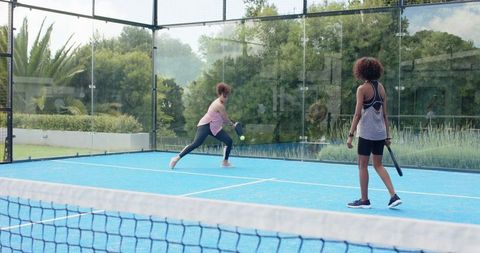 African american women playing padel on blue court with glass walls, training rally duo