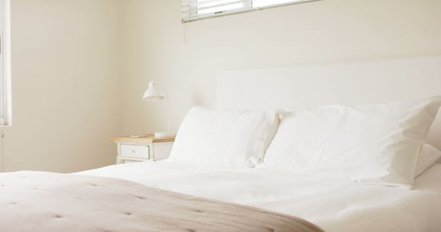 This image depicts a minimalistic white bedroom featuring a comfortable bed with white sheets and pillows, beside which stands a small white nightstand with a reading lamp. The soft, ambient lighting adds a peaceful and cozy atmosphere. This image is suitable for use in marketing materials for home decor, interior design inspiration, real estate listings, sleeping aids, and lifestyle blogs showcasing cozy living spaces.