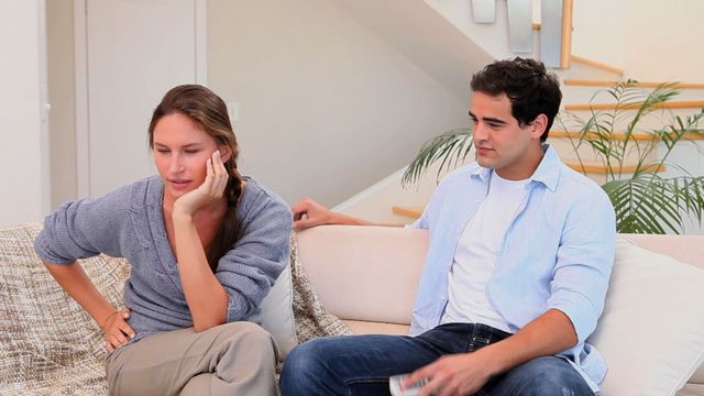 Couple sitting on a couch with woman looking upset while man attempts to engage. Useful for illustrating themes of relationship challenges and communication issues within domestic settings. Can be used in articles or content related to conflict resolution or emotional health.