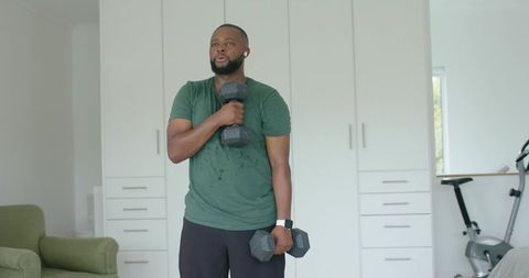 African american man lifting hex dumbbells in home gym wearing smartwatch and earbuds
