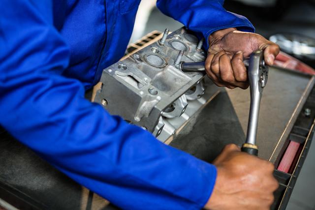 Mechanic in blue uniform focusing on repairing an engine part using a ratchet. Perfect for illustrating automotive repair services, mechanical engineering expertise, or showcasing hands-on skills in a workshop environment.