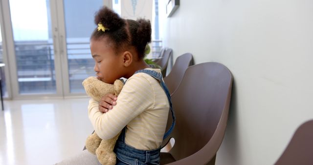 African american girl patient holding teddy bear sitting in waiting room in hospital. Medicine, healthcare and hospital, unaltered.