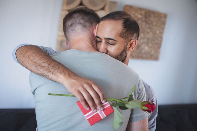 Diverse gay couple sharing a tender moment in their living room, with one holding a wrapped gift and a red rose. Ideal for use in articles or advertisements about LGBTQ relationships, romantic gestures, and celebrating love during quarantine. Can also be used for promoting inclusivity and diversity in relationships.