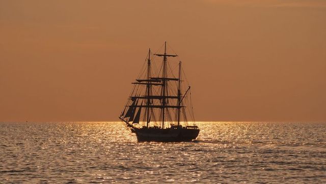 Three-masted ship sailing under orange sunset sky