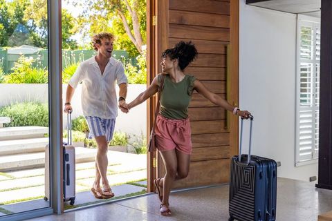 Diverse couple entering sunlit modern home holding hands and pulling suitcases