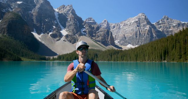 Male wearing sunglasses and life jacket paddling canoe on turquoise lake surrounded by mountains. Perfect for promotions of outdoor adventures, nature exploration, athletic lifestyle, travel and tourism advertisements, and recreational activities.