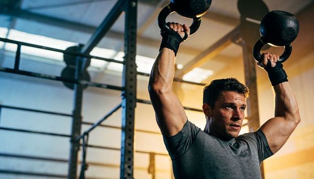 Athletic man pressing kettlebells overhead in industrial gym, strength training, focused