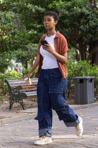 African woman walking through urban park holding smartphone and takeaway coffee, casual street style