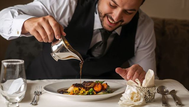 Smiling man in waistcoat pouring sauce over plated gourmet dish during elegant fine dining
