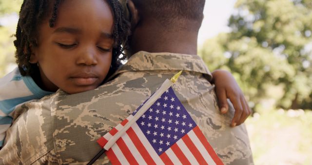 Child seen hugging parent dressed in military uniform, holding small American flag outdoors. Image reflects themes of family, love, patriotism, homecoming, and military service. Ideal for use in campaigns related to Veterans Day, Memorial Day, military family support, and patriotic events. Evokes strong emotional connection, highlighting support and bond within military families.