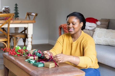 African american woman wrapping holiday gifts at home wearing mustard sweater in cozy living room