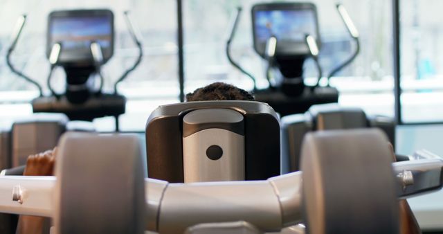This image captures a person working out on a weight machine in a gym. The focus is set on the machine and their back. In the background, there are blurred treadmills equipped with screens. Ideal for use in fitness blogs, exercise tutorials, health and wellness promotions, or gym advertisements aiming to highlight modern equipment and a motivating environment.