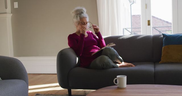 This image depicts an elderly woman sitting on a sofa reading a book and holding glasses. There is a coffee mug on a table in the foreground, and natural light is coming through windows, creating a cozy atmosphere. This can be used for themes related to retirement, leisure activities, lifestyle, and home comfort.