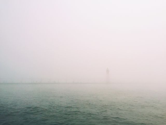 Image shows a lighthouse barely visible through dense fog over a tranquil sea. Perfect for concepts of mystery, serenity, and maritime themes. Can be used in articles, blogs, or marketing materials related to weather, seafaring, navigation, and natural phenomena.