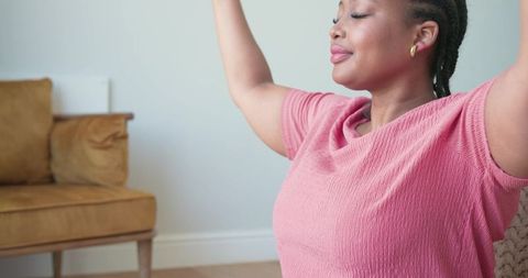 African american woman stretching arms overhead at home in pink top, minimalist living room