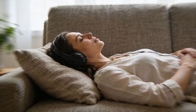 Tranquil woman listening to music on headphones while resting on sunlit grey sofa at home