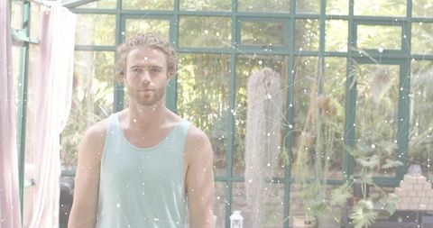 Young man standing in sunlit glasshouse wearing teal tank with botanical network overlay