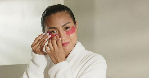 Mid-adult asian woman adjusting under-eye gel patches in white robe for skincare routine