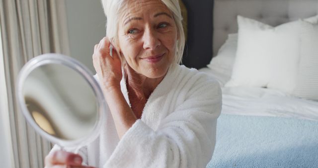 Senior woman wearing a white bathrobe holding hand mirror while in bedroom, focusing on personal grooming and self-care. Portrays themes of aging gracefully, morning routine, and tranquility. Ideal for advertisements, blogs, or articles related to senior lifestyle, beauty, and self-care products.