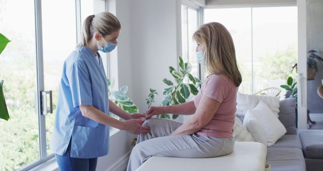 Home nurse in medical mask assisting elderly woman with leg physical therapy exercises in residential setting. Suitable for topics related to healthcare, senior care, rehabilitation, home medical services, and patient wellbeing.