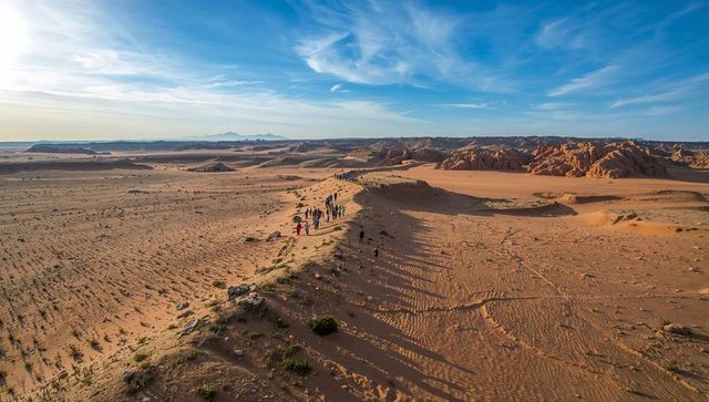 Hikers crossing dune ridge under expansive blue sky at golden-hour desert trek with backpacks