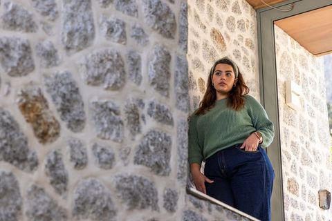 Woman leaning on stone wall at doorway wearing green sweater and jeans, casual lifestyle