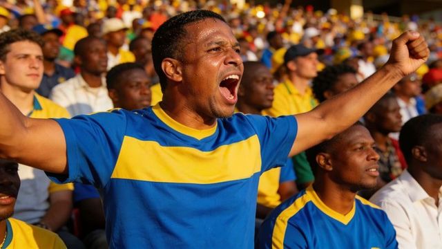 Passionate fan cheering in sunny stadium wearing blue yellow jersey and raising arms