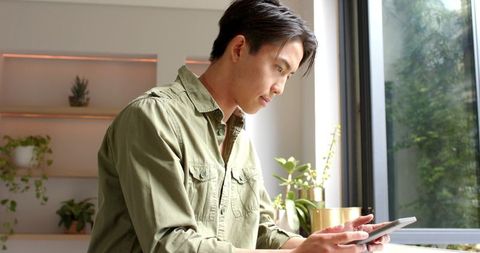 Young Asian Man Using Tablet by Sunny Windowsill with Plants
