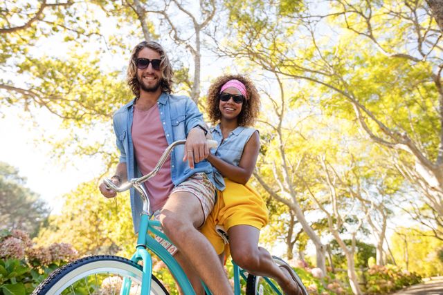 This image shows a happy couple cycling together in a park on a sunny day. They are both wearing casual summer clothing and sunglasses, enjoying the outdoor activity amidst lush greenery. This image can be used for promoting healthy lifestyles, outdoor activities, summer fun, and leisure time with loved ones.