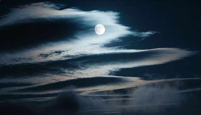 Full moon glowing through layered clouds and rippling bands over midnight sky with lunar glow