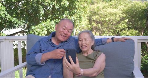 Senior asian couple sharing smartphone on porch smiling and pointing while sitting closely outdoors