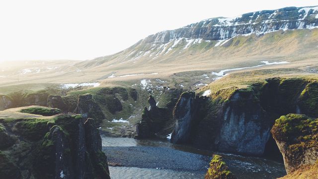 Scenic view of a rugged rocky canyon with a flowing river, captured at sunrise. Snow patches dot the highland and grassy cliffs rise from the valley below. Perfect for use in travel blogs, environmental presentations, or nature-themed posters. Highlights the majestic beauty and raw wilderness of Iceland's landscapes.
