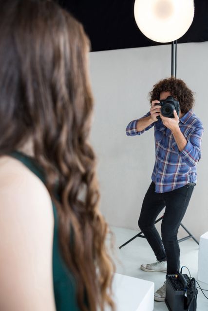 Female model posing for a professional photoshoot in a studio setting. Photographer with curly hair and plaid shirt capturing the moment with a camera. Ideal for use in articles or advertisements related to fashion, photography, modeling, and studio setups.