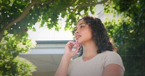 Sunlit woman standing under leafy tree, hand on chin, serene contemplative portrait