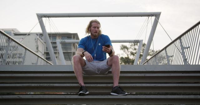 Young man sitting on bridge steps, using smartphone and listening to music with earphones. Ideal for illustrating urban lifestyle, fitness, relaxation, or technology in everyday life.