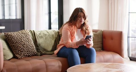 Concerned woman sitting on tan leather sofa checking smartphone in bright living room