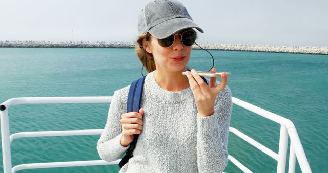 Woman speaking on smartphone with ocean background. She is wearing a casual sweater, a hat, and sunglasses, with a backpack. Ideal for travel, tourism, and technology websites, promotional materials, and lifestyle blogs. Captures relaxation, modern communication, and outdoor adventure concepts.