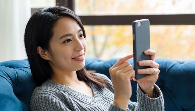 Young woman relaxing on cozy blue sofa scrolling smartphone by window with autumn bokeh