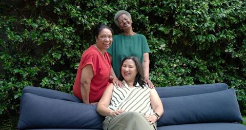 Diverse older women laughing on outdoor sofa in garden, sharing friendship and joy