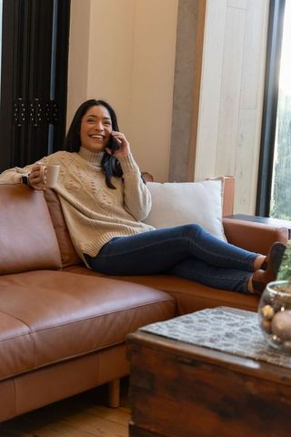 Smiling asian woman relaxing on leather sofa, talking on smartphone and sipping coffee