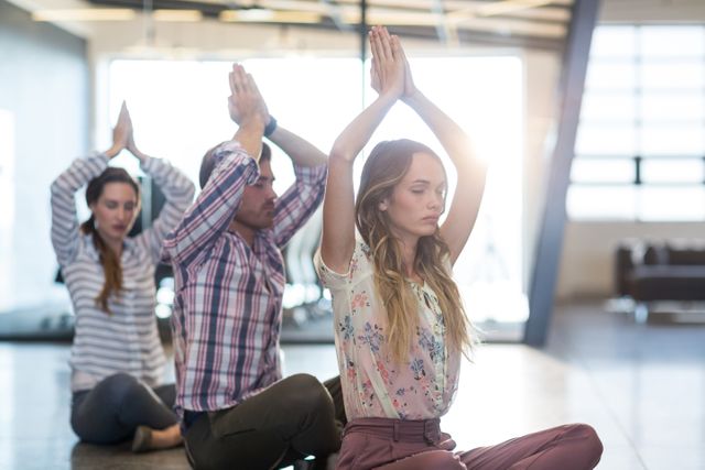 Business professionals practicing yoga in an office setting. They are sitting on the floor with their hands raised above their heads in a meditative pose. This image can be used for promoting corporate wellness programs, stress relief techniques, team building activities, and healthy lifestyle initiatives in the workplace.