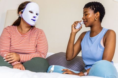African american friends using led facial mask and handheld skincare device at home