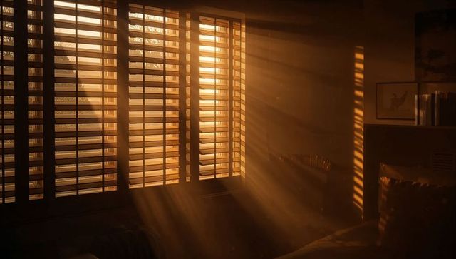 Golden Sunlight Streaming Through Wooden Shutters Casting Dust Rays Across Cozy Bedroom