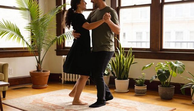 Couple dancing barefoot on rug in sunlit living room with lush houseplants